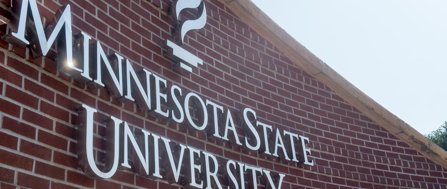 Close-up view of the metal Minnesota State University, Mankato logo that's attached to the brick entrance sign. The sun reflects off the metal.